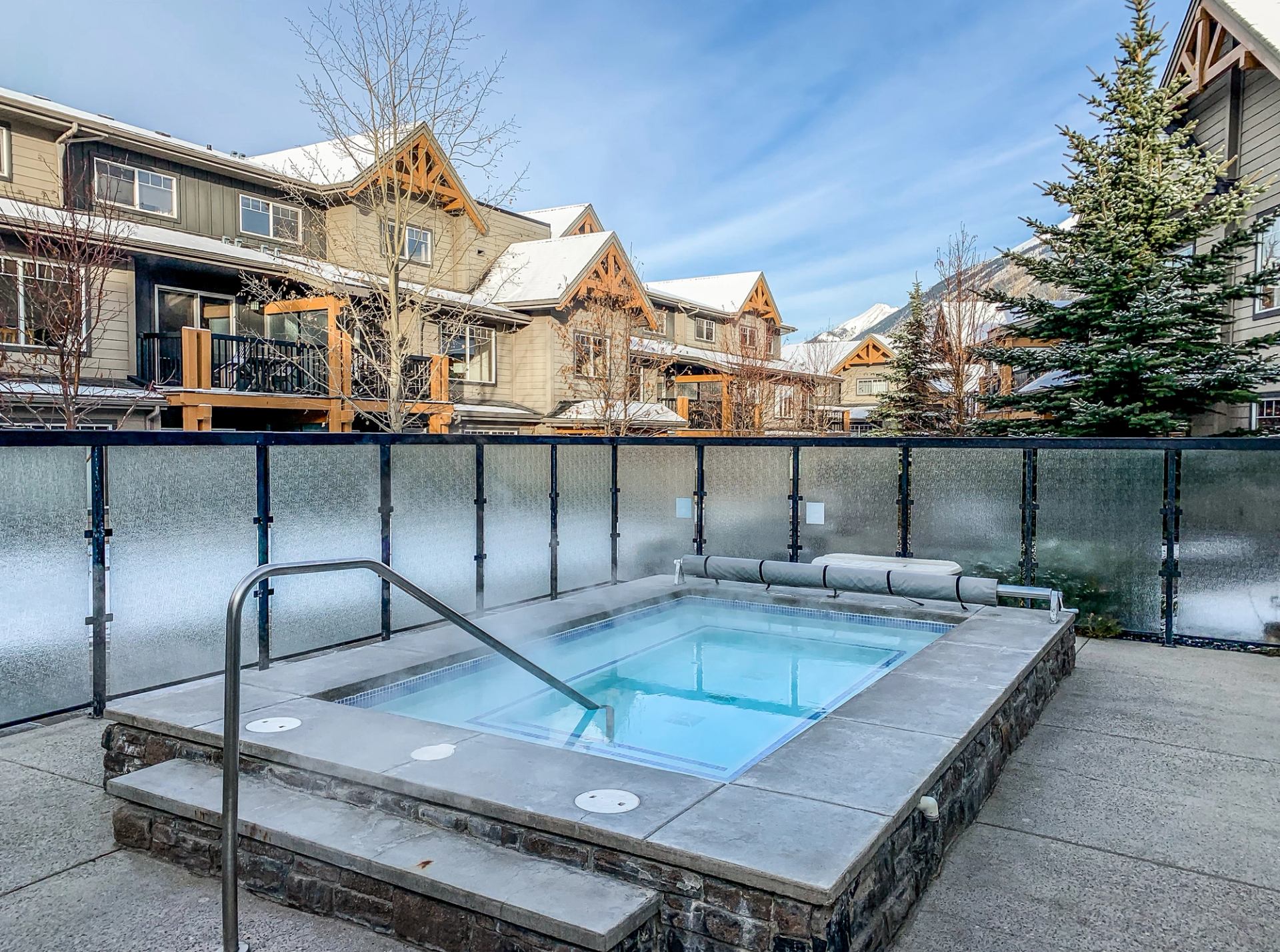 Steaming outdoor hot tub on a patio in front of snow-covered buildings and trees.