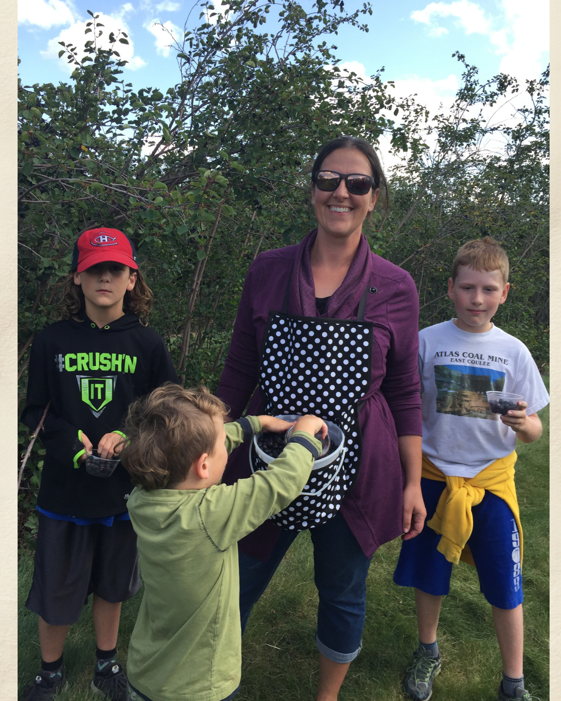 Children and adult stand outdoors near bushes while placing items into a polka-dot bag.