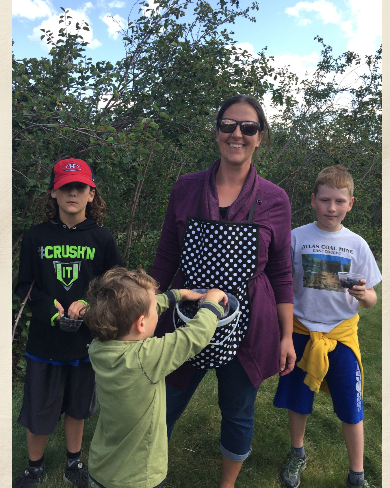 Children and adult stand outdoors near bushes while placing items into a polka-dot bag.