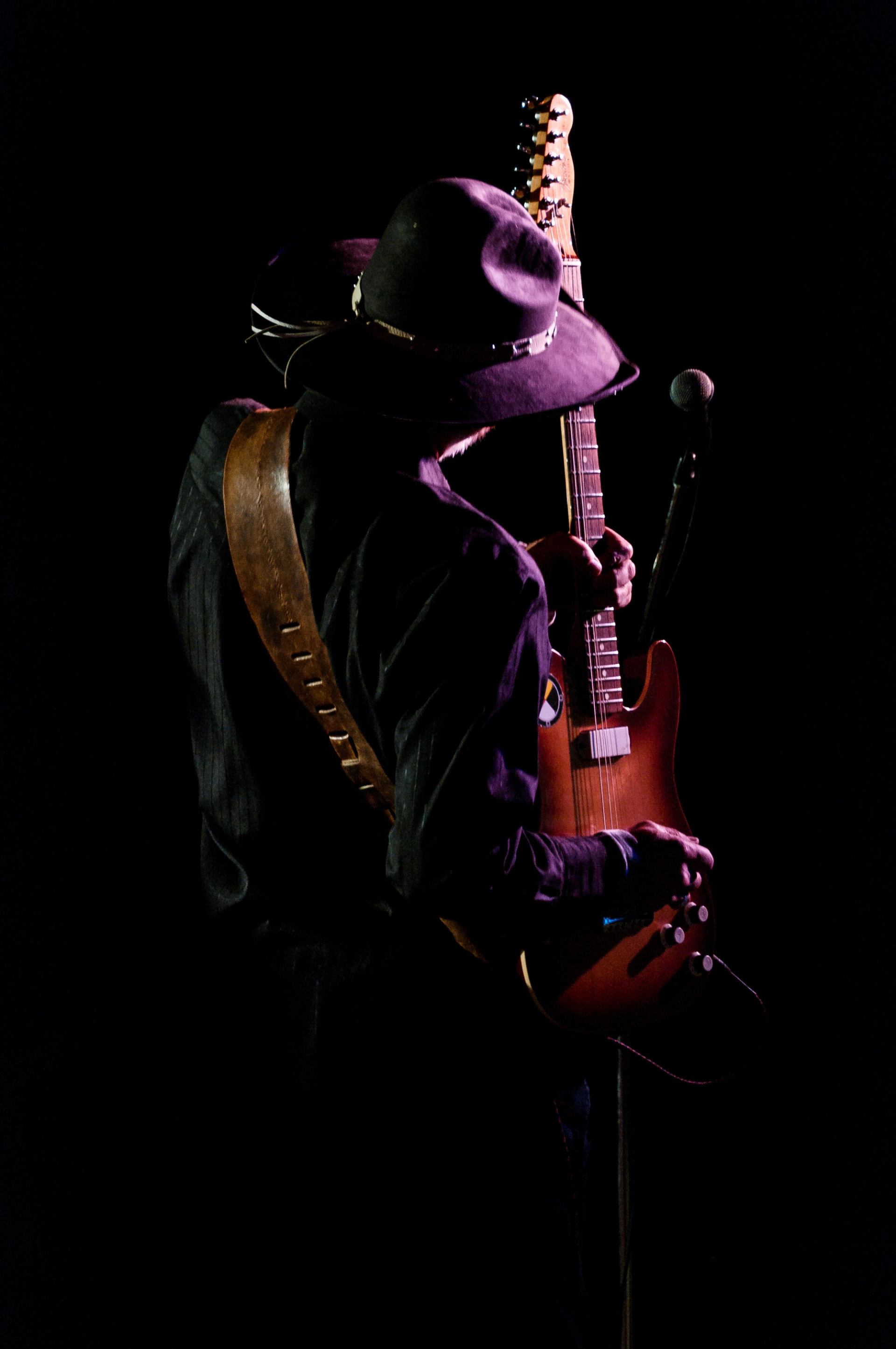 Person in cowboy hat holds electric guitar beside microphone on dark stage.