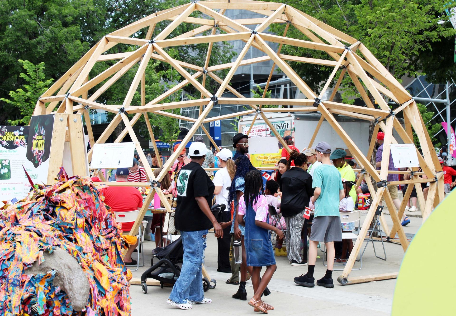 Outdoor festival crowd gathers under wooden geodesic dome with activity tables and posters.
