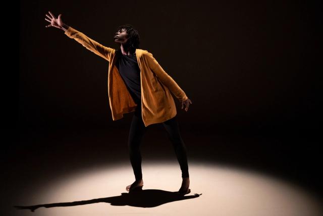 A barefoot performer stretches their hand upwards, bathed in the glow of a spotlight on stage at the Fringe Theatre Arts Barns