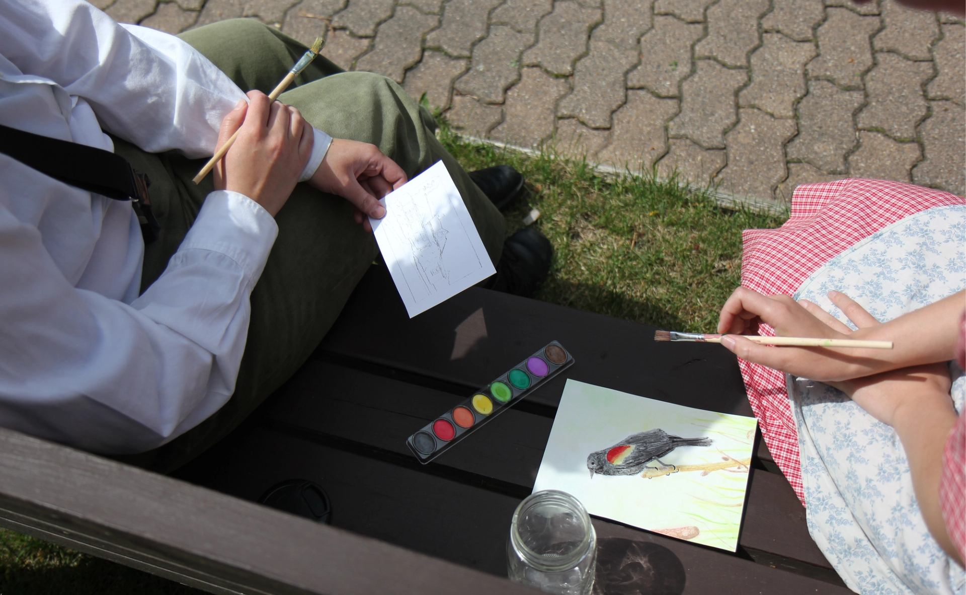 Hands holding brushes over watercolour set and bird painting on outdoor bench.