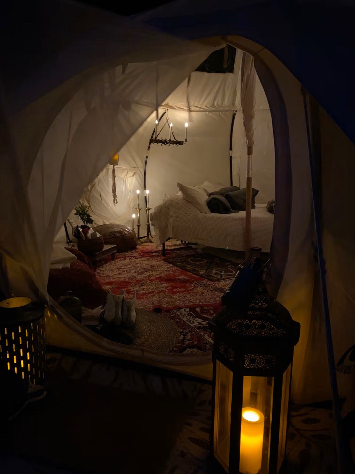 Glamping tent interior with lit lanterns, bed, and patterned rug near a hanging chandelier.