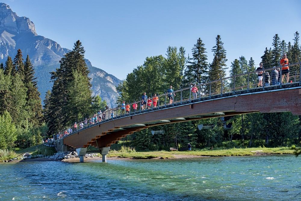 Large group running across a bridge over a river with mountains behind.