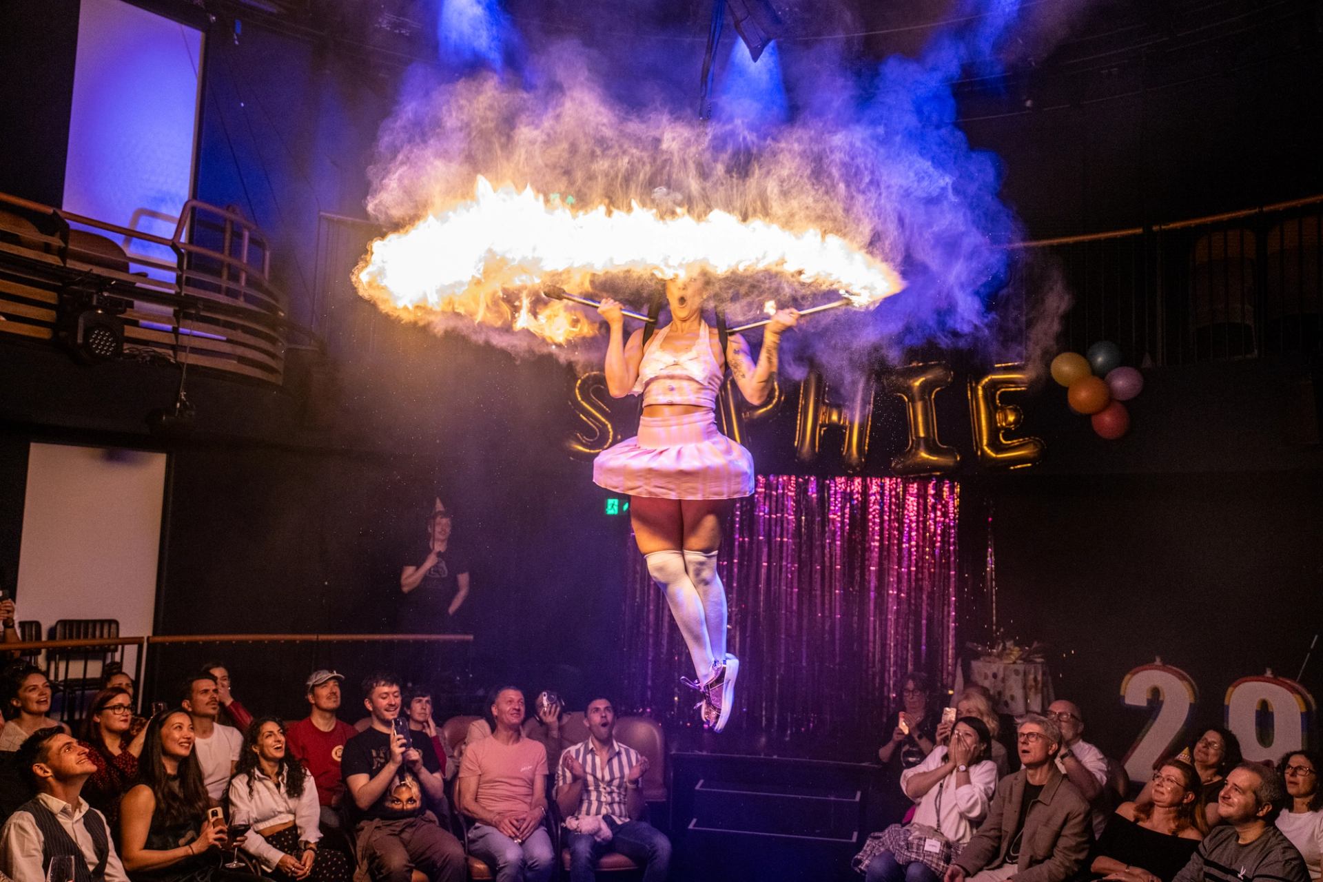 Performer in pink costume spins flaming hoop above audience in decorated theatre.