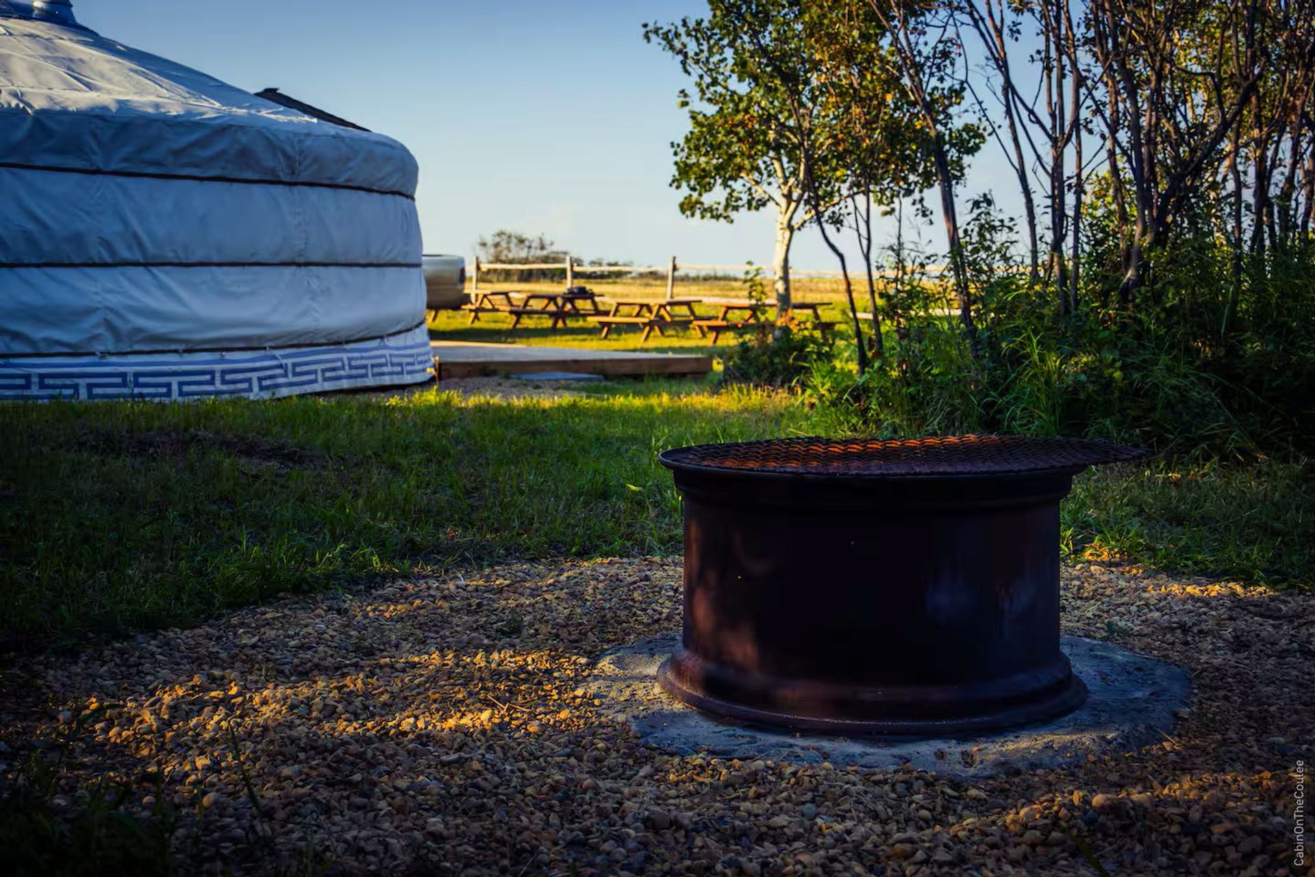 Metal fire pit near a white yurt with trees and picnic tables in the background.