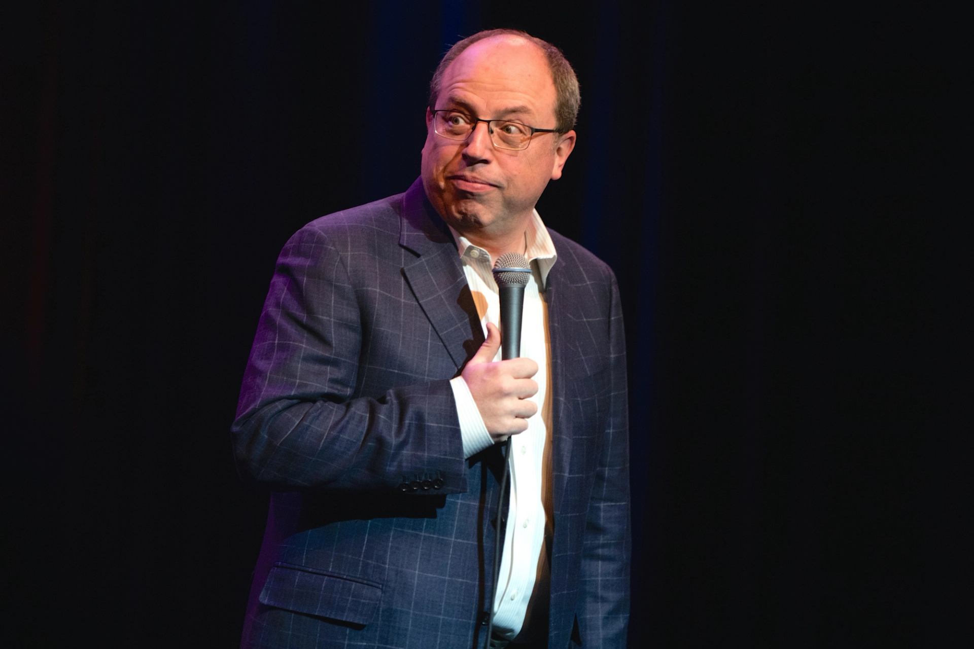 Comedian on stage holds a microphone and looks sideways against dark background.