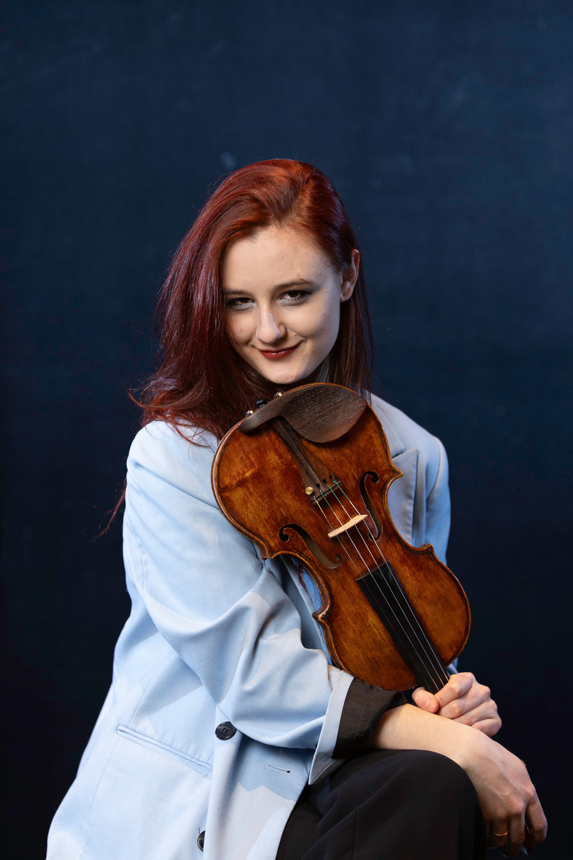 Red-haired violinist in a light blue blazer holds a violin against a dark backdrop.