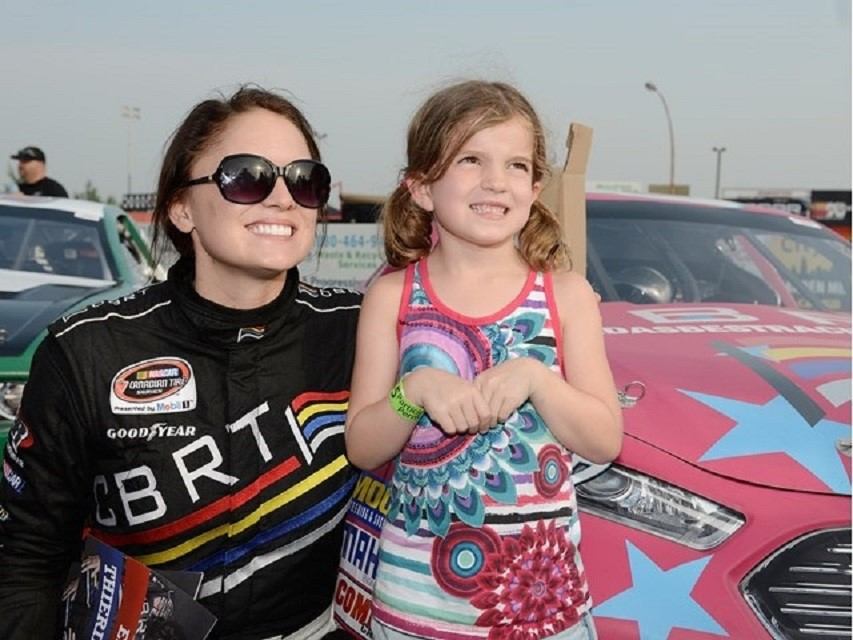 Person in a racing suit standing with a child in front of a brightly decorated NASCAR car.