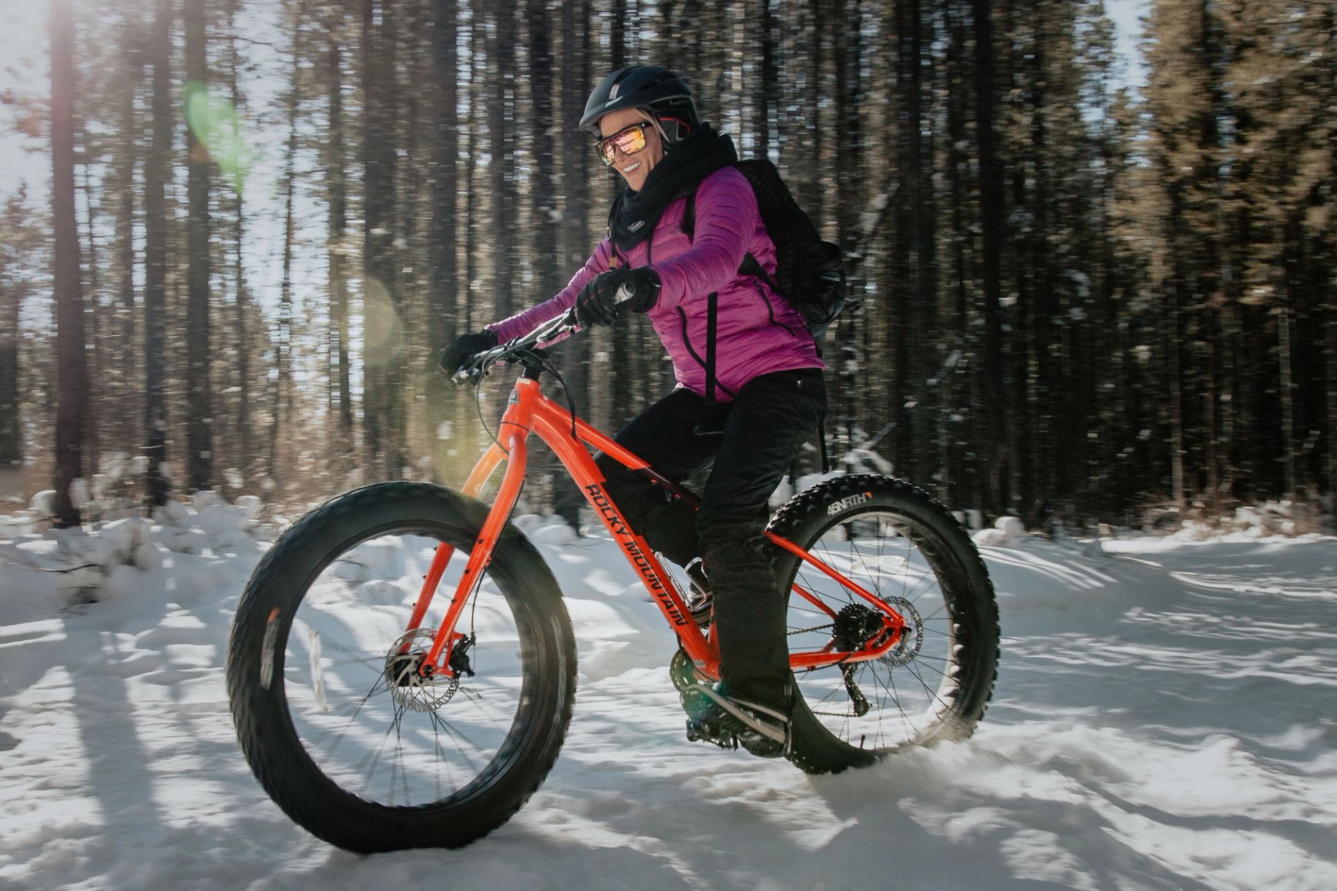 Person rides orange fat-tire bike on snowy forest trail in winter gear.