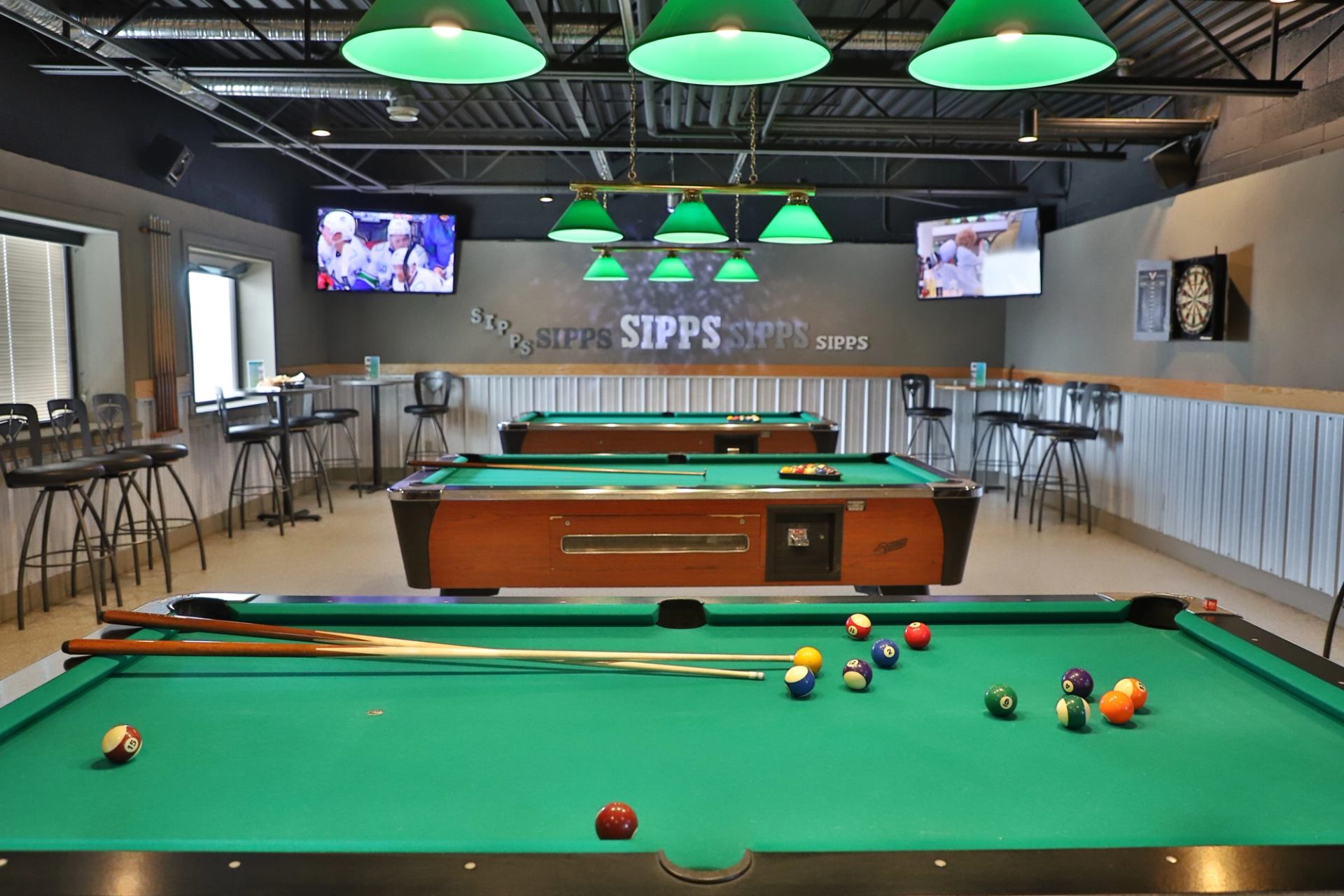 Pool tables with cues and balls under green lights, SIPPS wall sign and TVs.