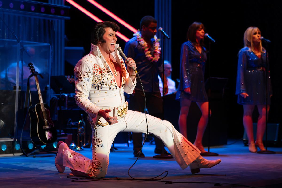 Elvis impersonator in white jumpsuit kneels singing on stage with backup singers.