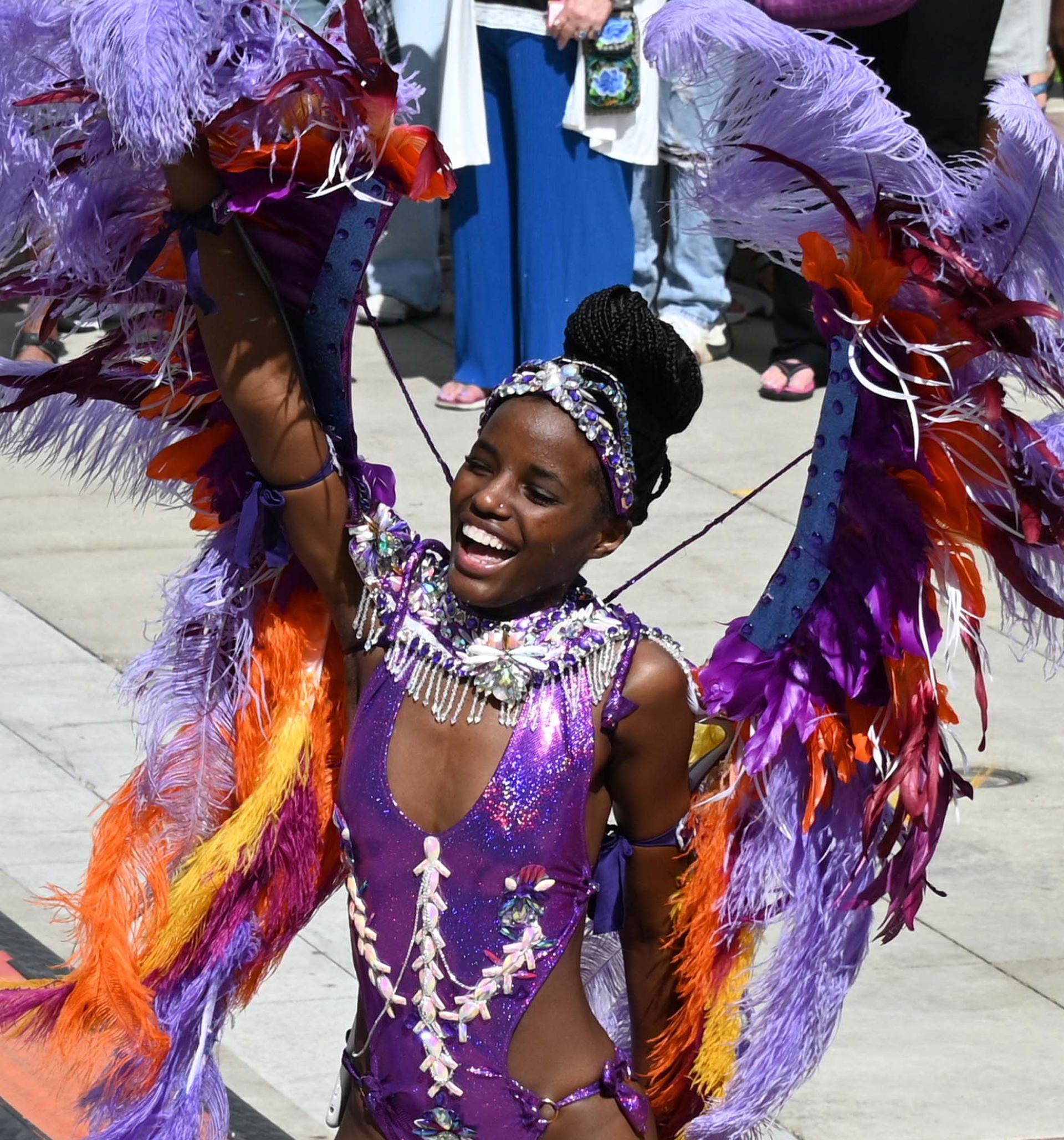 Carnival performer in purple feathered costume raises arm while walking past onlookers.