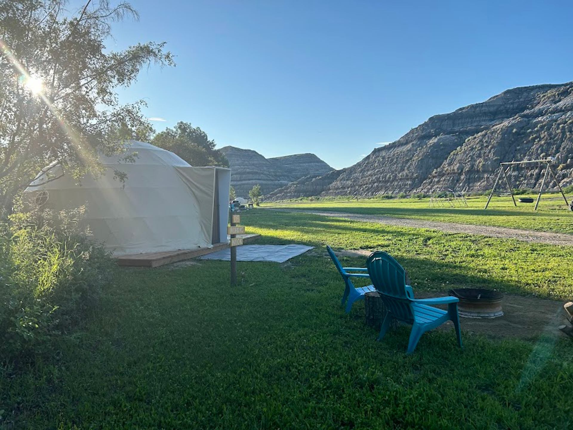 Yurt beside grassy campsite with blue chairs, fire pit and swing set near hills.