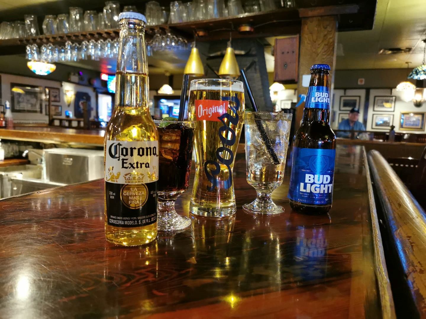 A bar setting with three drinks: a Corona Extra beer, a glass of cola with ice, and a Coors glass with water and a straw. A Bud Light bottle is beside them. The atmosphere is warm and inviting.