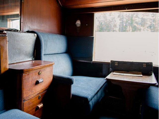 Cozy vintage train cabin with wooden walls, blue upholstered seats, and a small dining table by the window. A wooden drawer unit adds a classic touch.