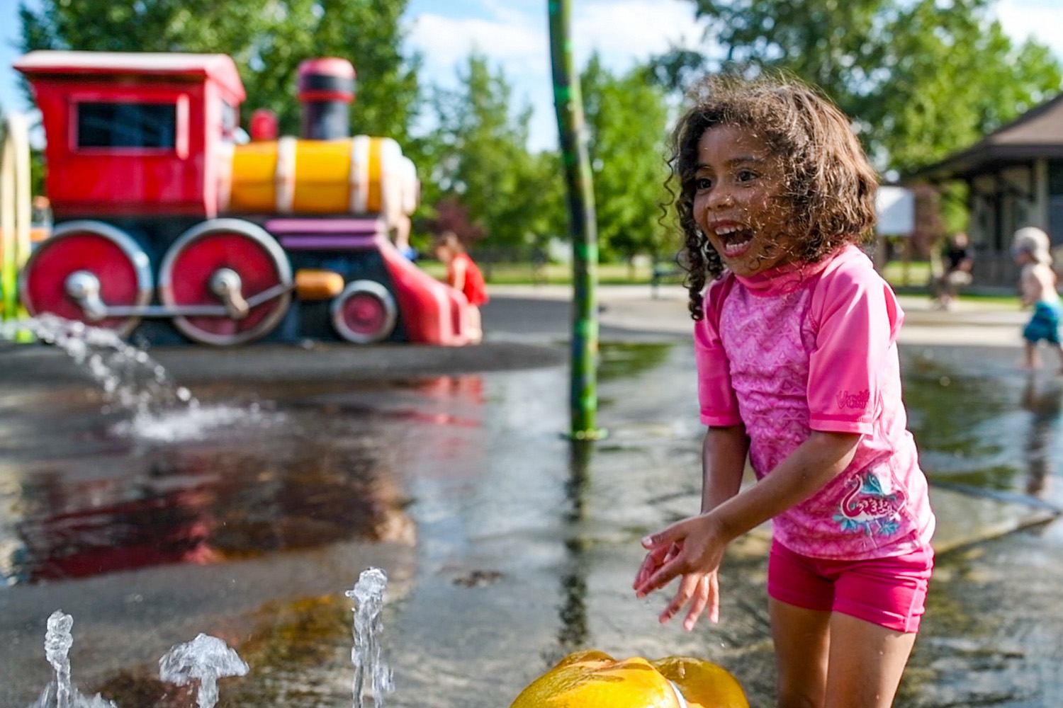 Child in pink plays near water jets at a colorful outdoor splash park with train structure.