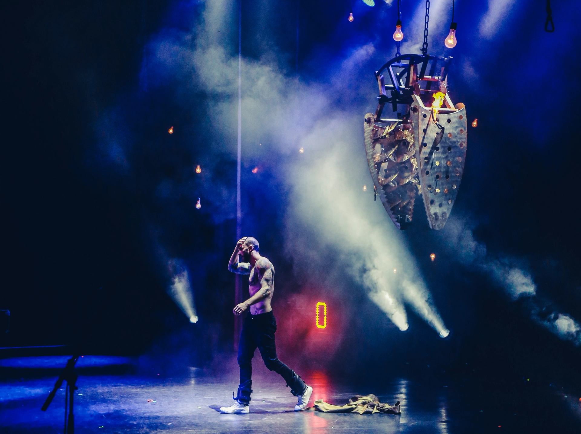 Stage performer walks shirtless past hanging metal structure amid blue smoke and spotlights.