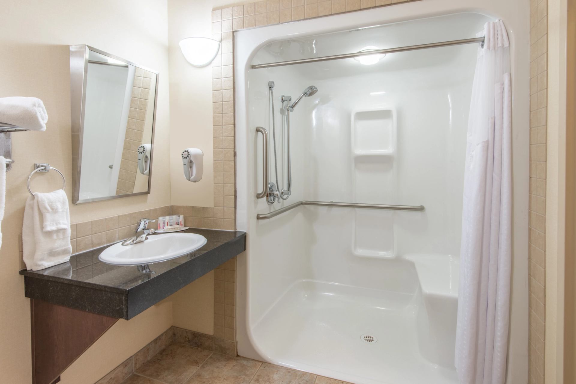 Accessible hotel bathroom with roll-in shower, grab bars, sink, mirror and towels.