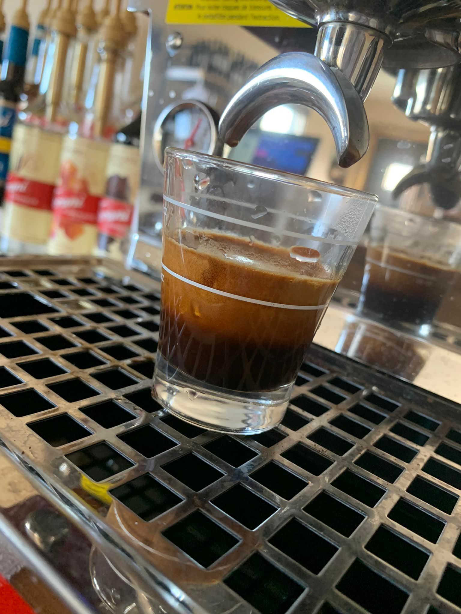 Glass cup of dark coffee on a metal counter, surrounded by other drinks in focus.