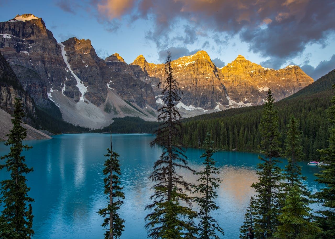 Turquoise mountain lake with evergreen forest and rocky peaks under partly cloudy sky.