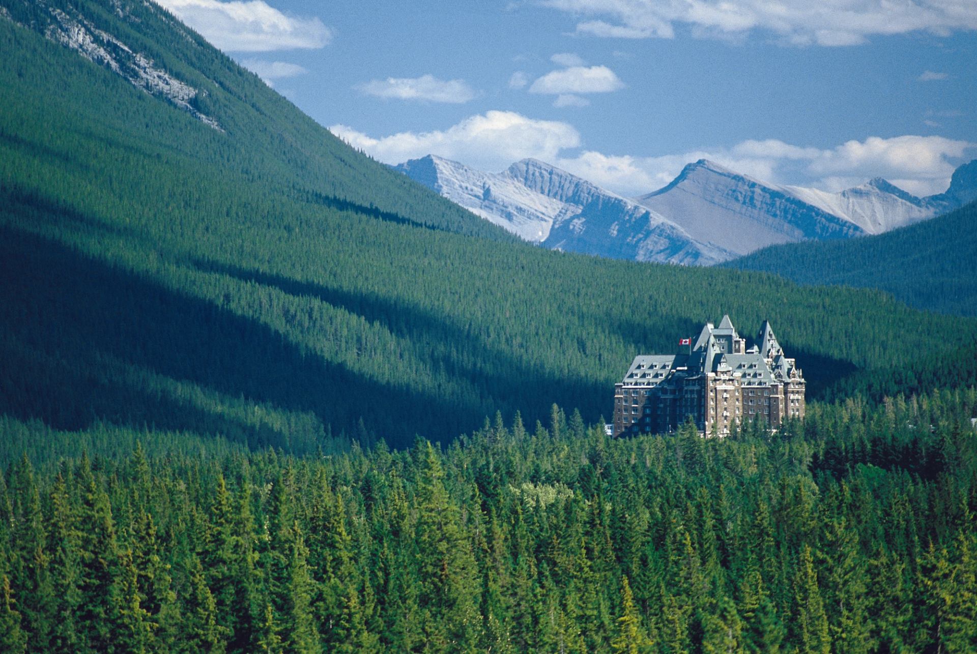 Castle-like hotel with Canadian flag surrounded by dense evergreen forest and mountains.