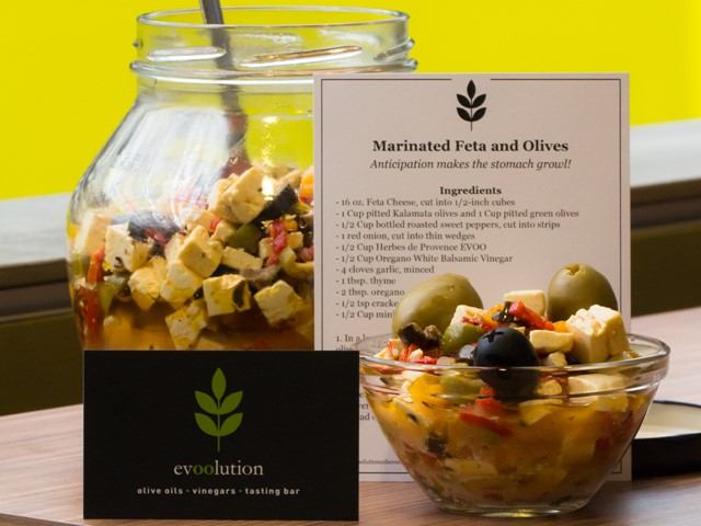 Glass jar of marinated feta and olives with recipe card on table, evoolution sign visible.