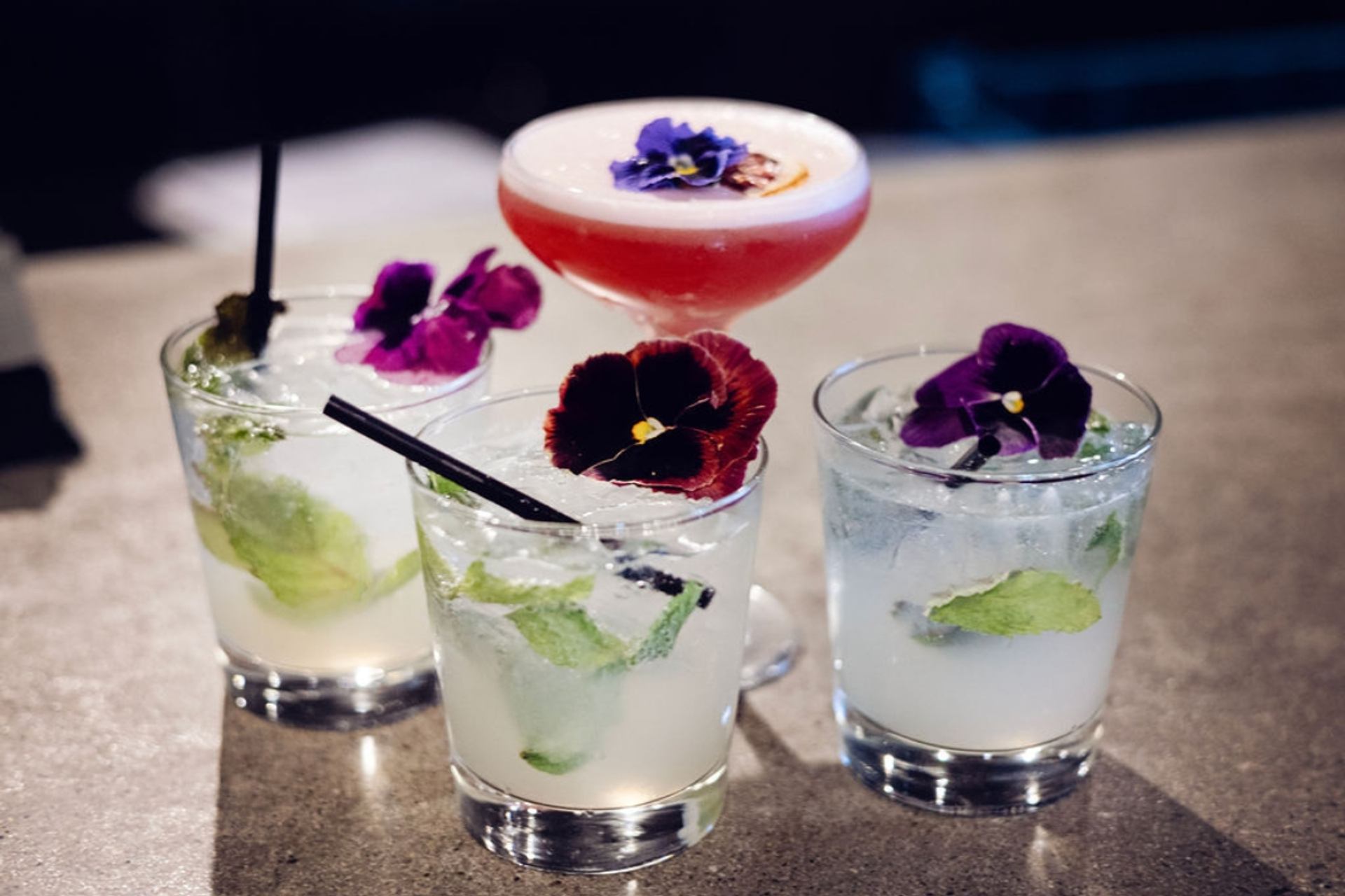 Four colourful cocktails garnished with flowers on a bar counter.