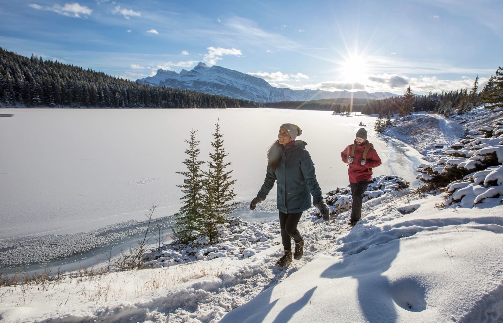 Two people hiking along a snowy lakeshore with mountains in the distance.