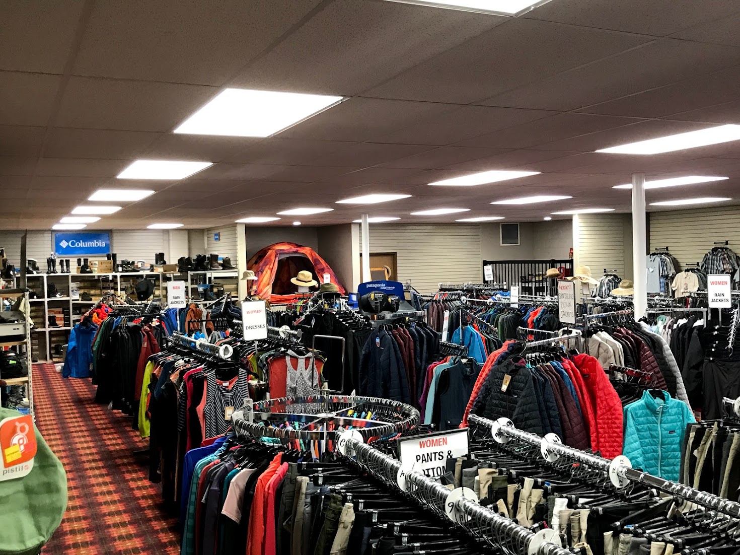 Clothing store interior with racks of jackets and shirts, camping gear shelves in background.