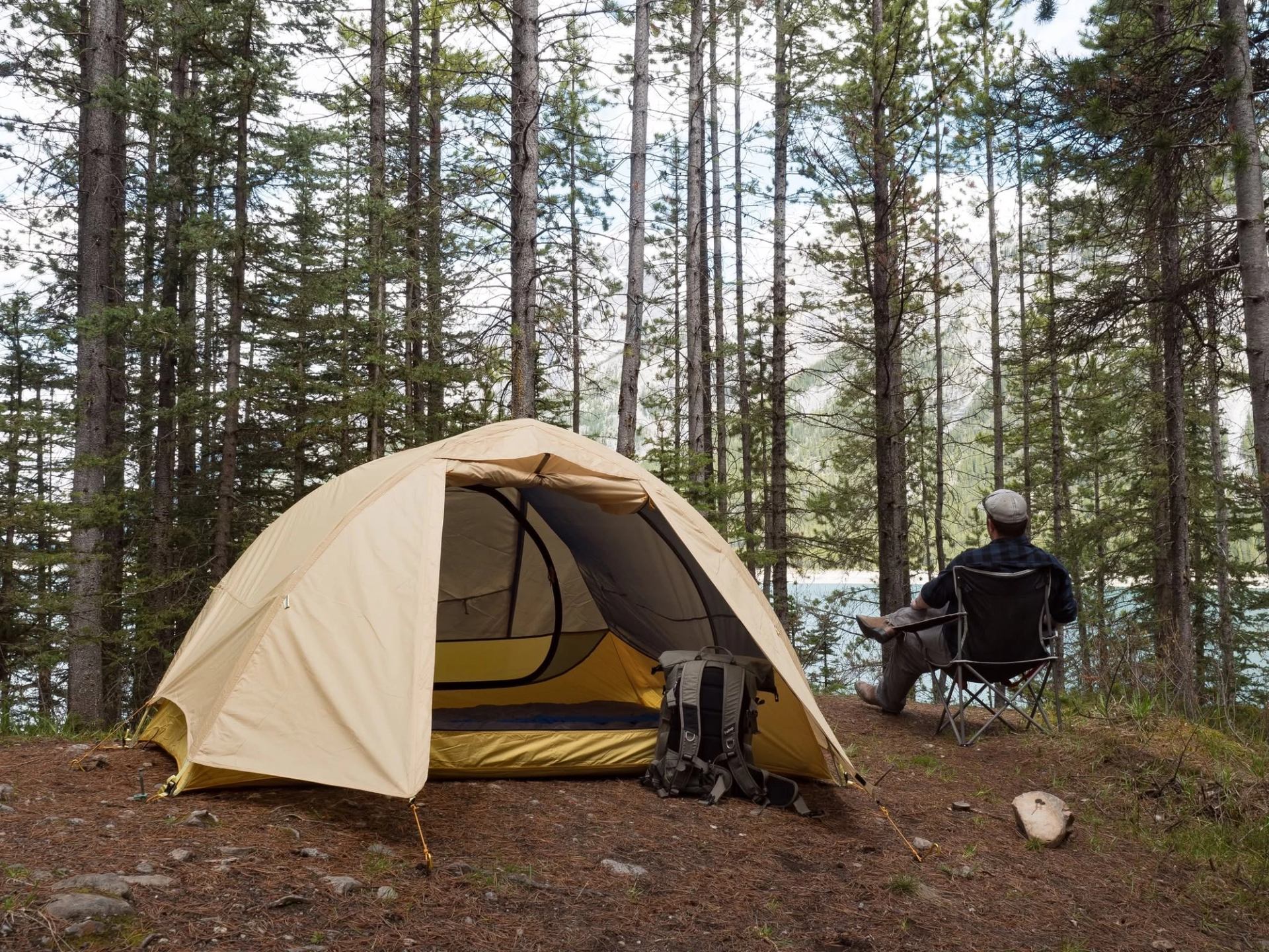 Camper sits beside a tent and backpack in a pine forest overlooking a lake.