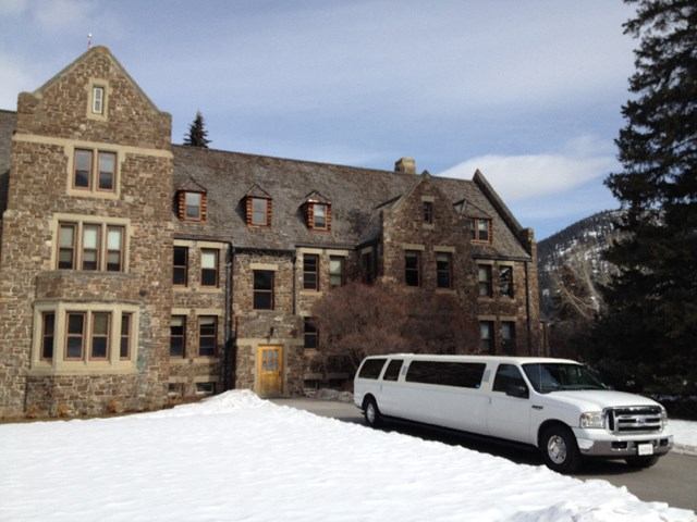White limo parked by stone building with snow, trees, and mountains in background.