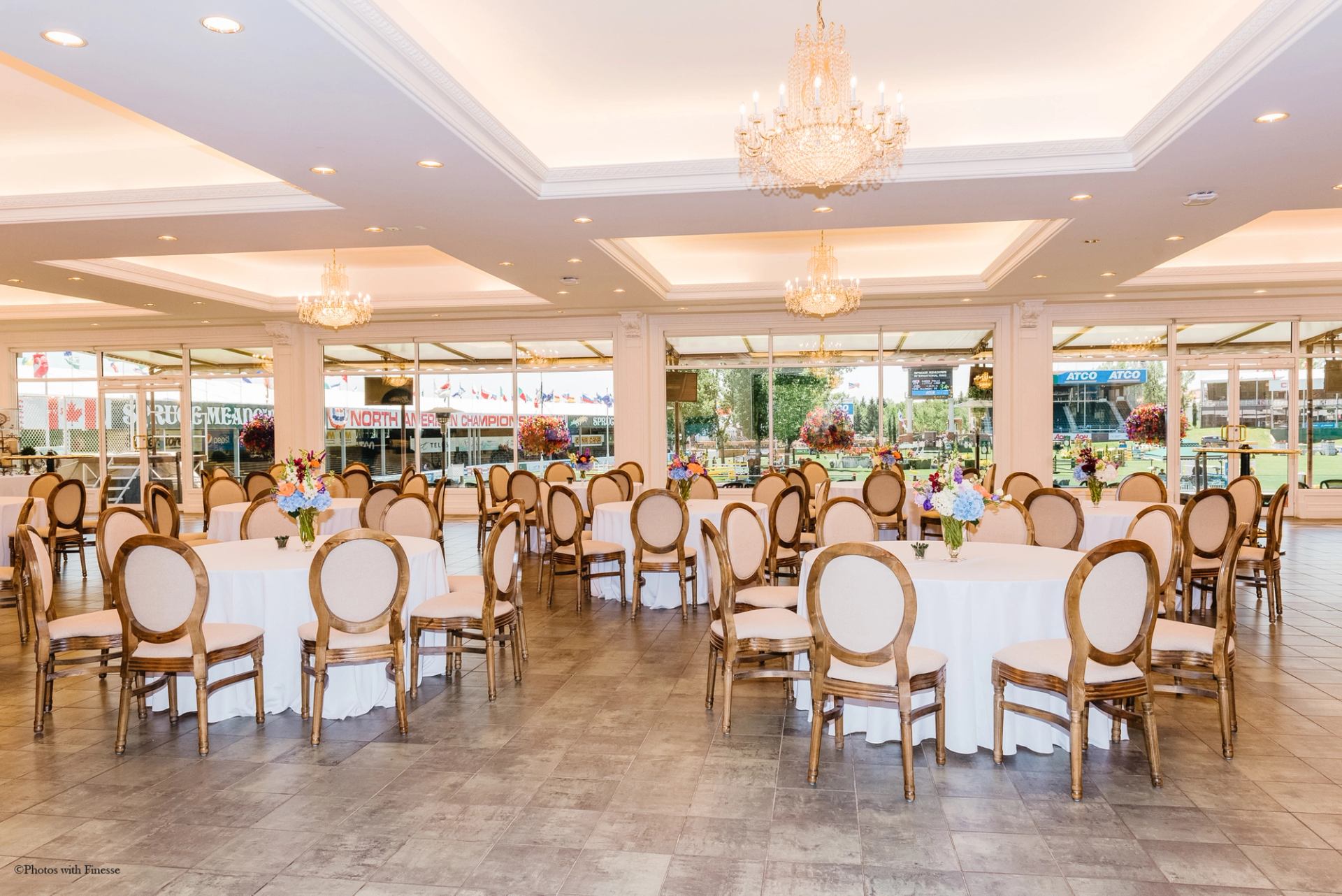 Bright banquet hall at Spruce Meadows with round tables, floral centerpieces, chandeliers, and large windows.