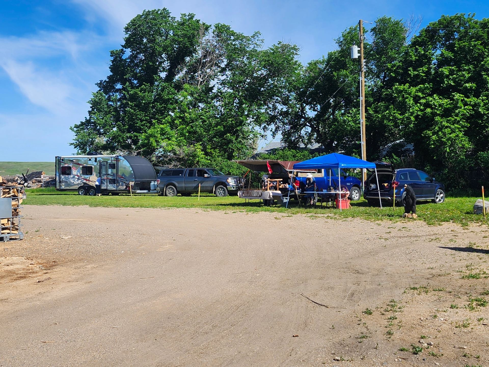 Outdoor campsite with trailer, trucks, blue canopy tent and people gathered near trees.