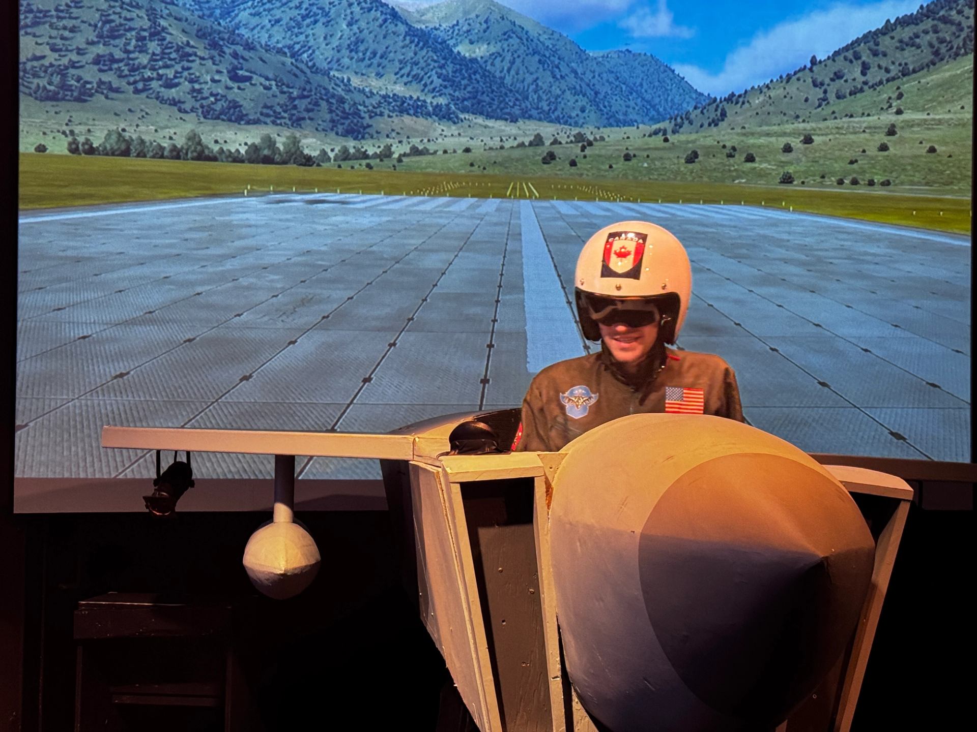 A person in a flight simulator cockpit with a runway and mountains on the screen backdrop.