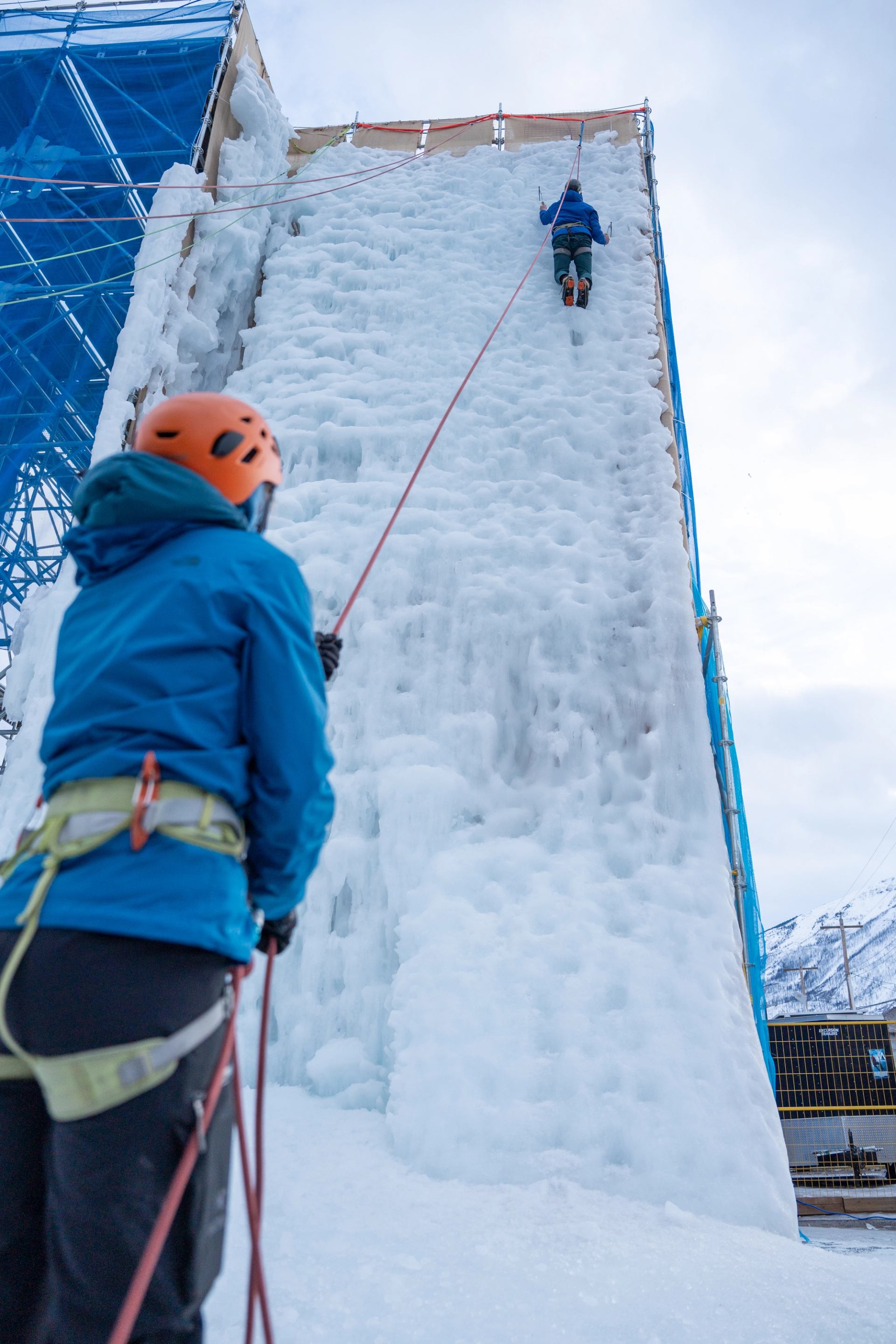 Two climbers on icy wall with ropes and gear beside blue-netted scaffolding.