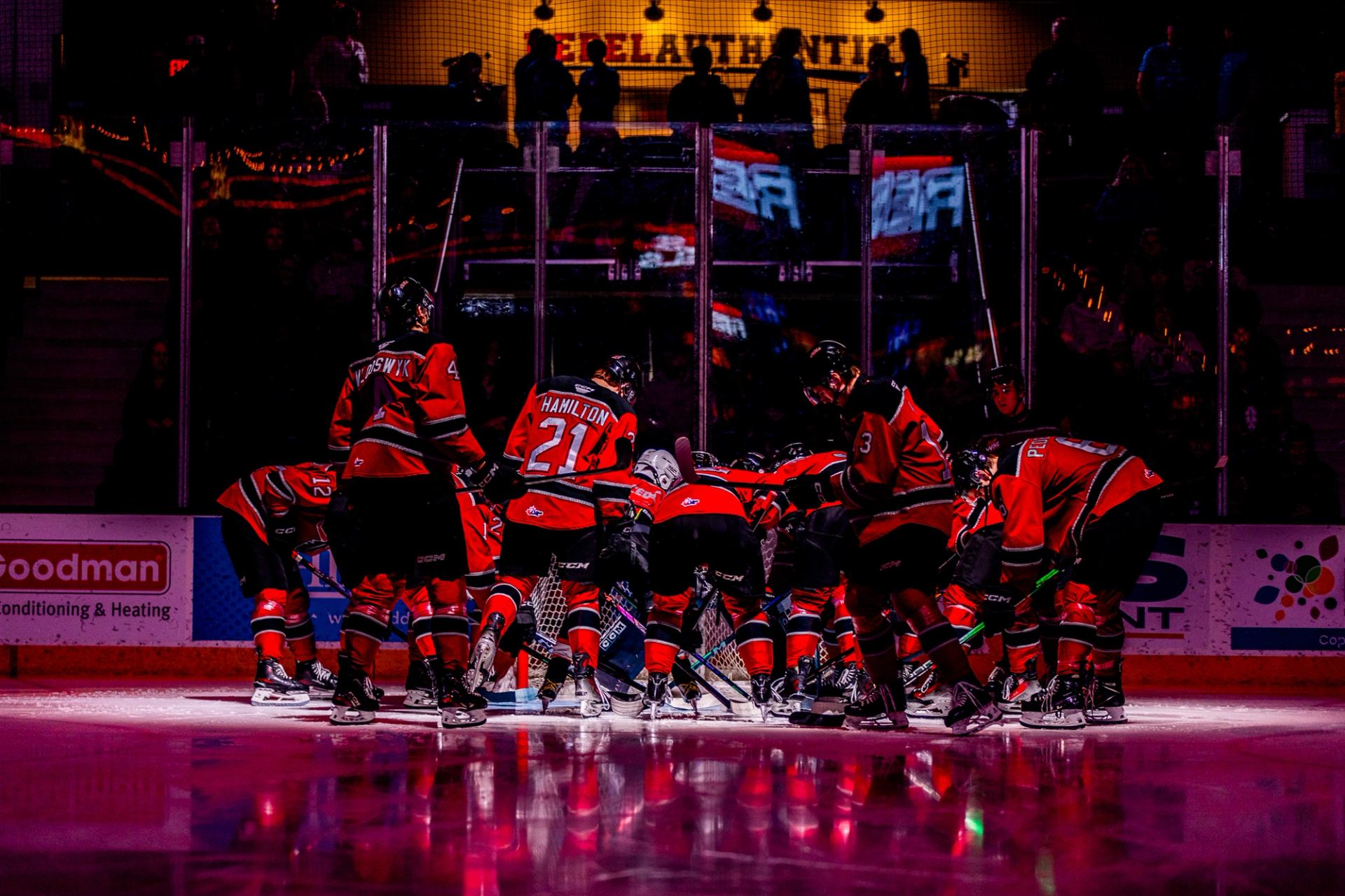 Red Deer Rebels players huddle together on the ice under dramatic arena lighting before a home game.
