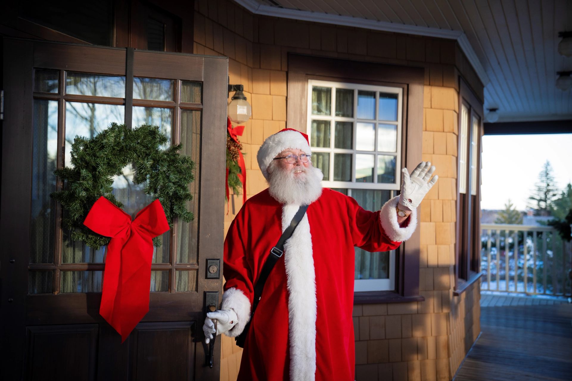 Santa waves cheerfully at the doorway decorated with a wreath and red bow.