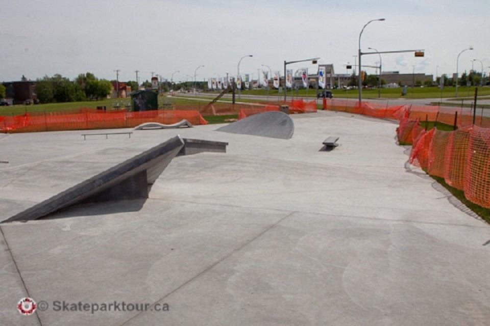 Concrete skatepark with ramps, rails, and a fenced perimeter along a walkway.