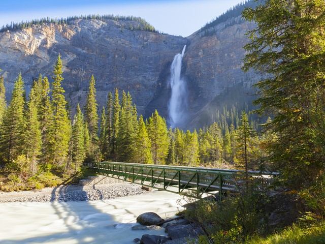 Waterfall with green footbridge over rocky river in forested mountain valley.