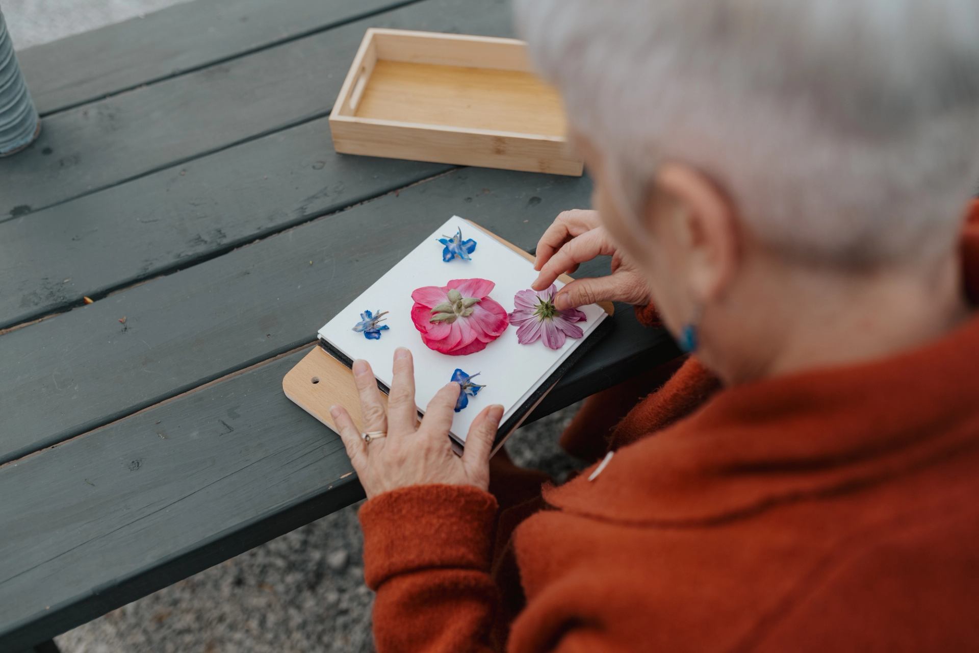 A person arranging pressed flowers on a small board at an outdoor table.