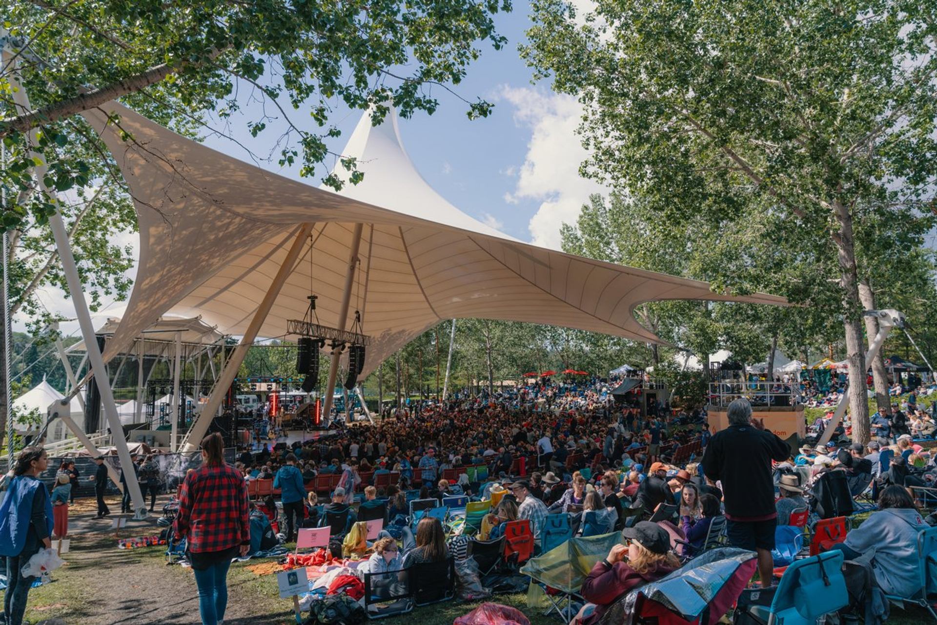 Outdoor music festival crowd under large white canopy, surrounded by trees and lawn chairs.