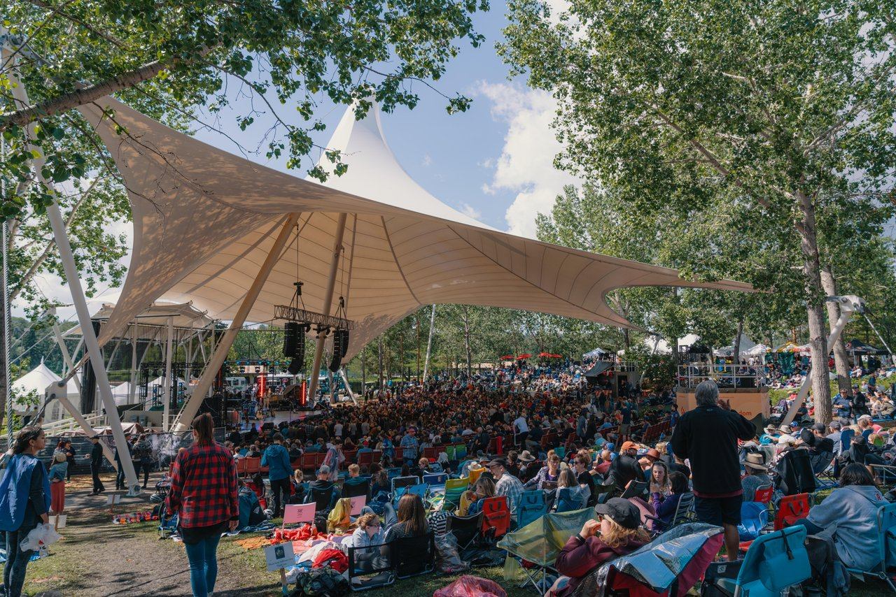 Outdoor music festival crowd under large white canopy, surrounded by trees and lawn chairs.