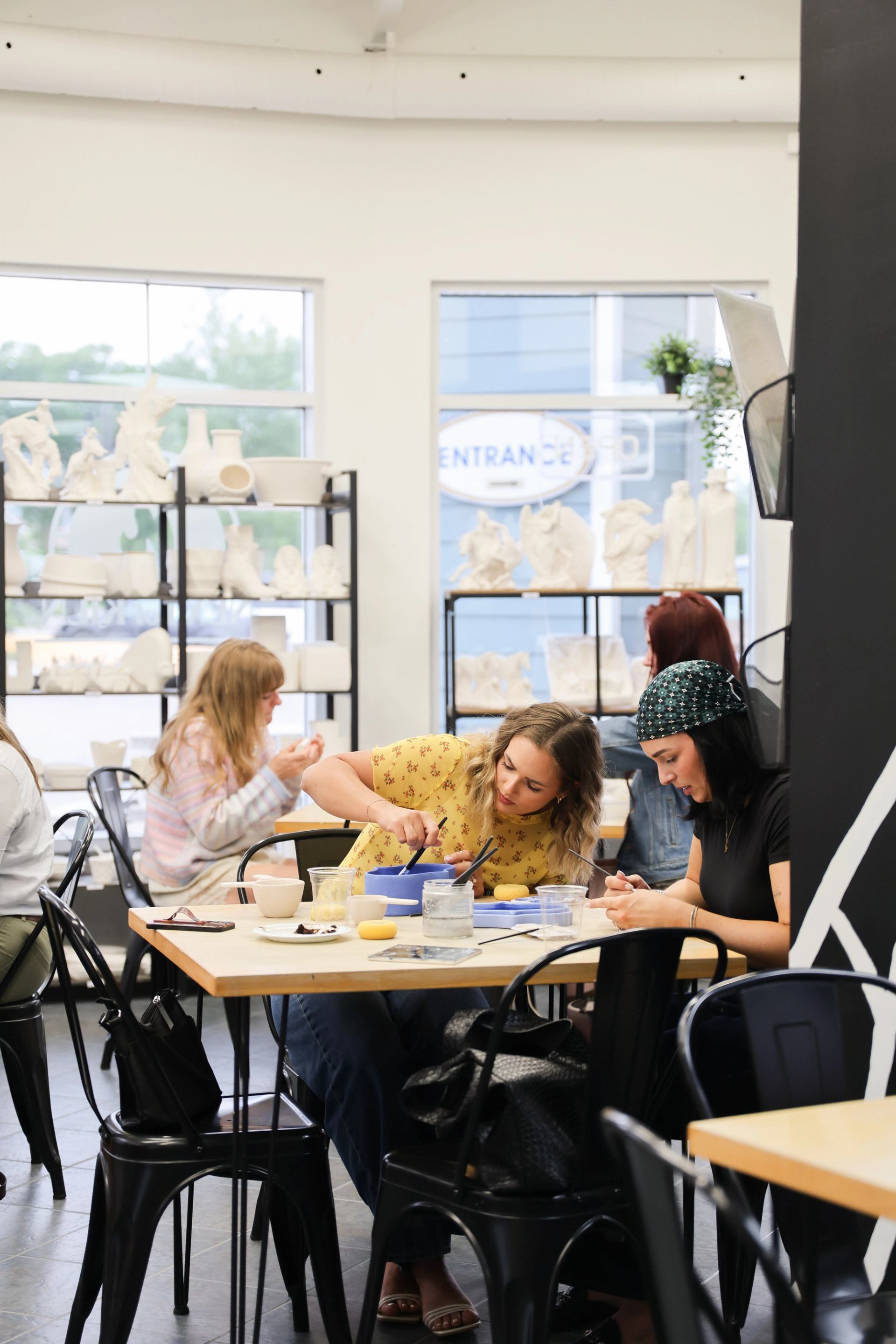 People painting pottery at tables in a bright studio with shelves of ceramics.