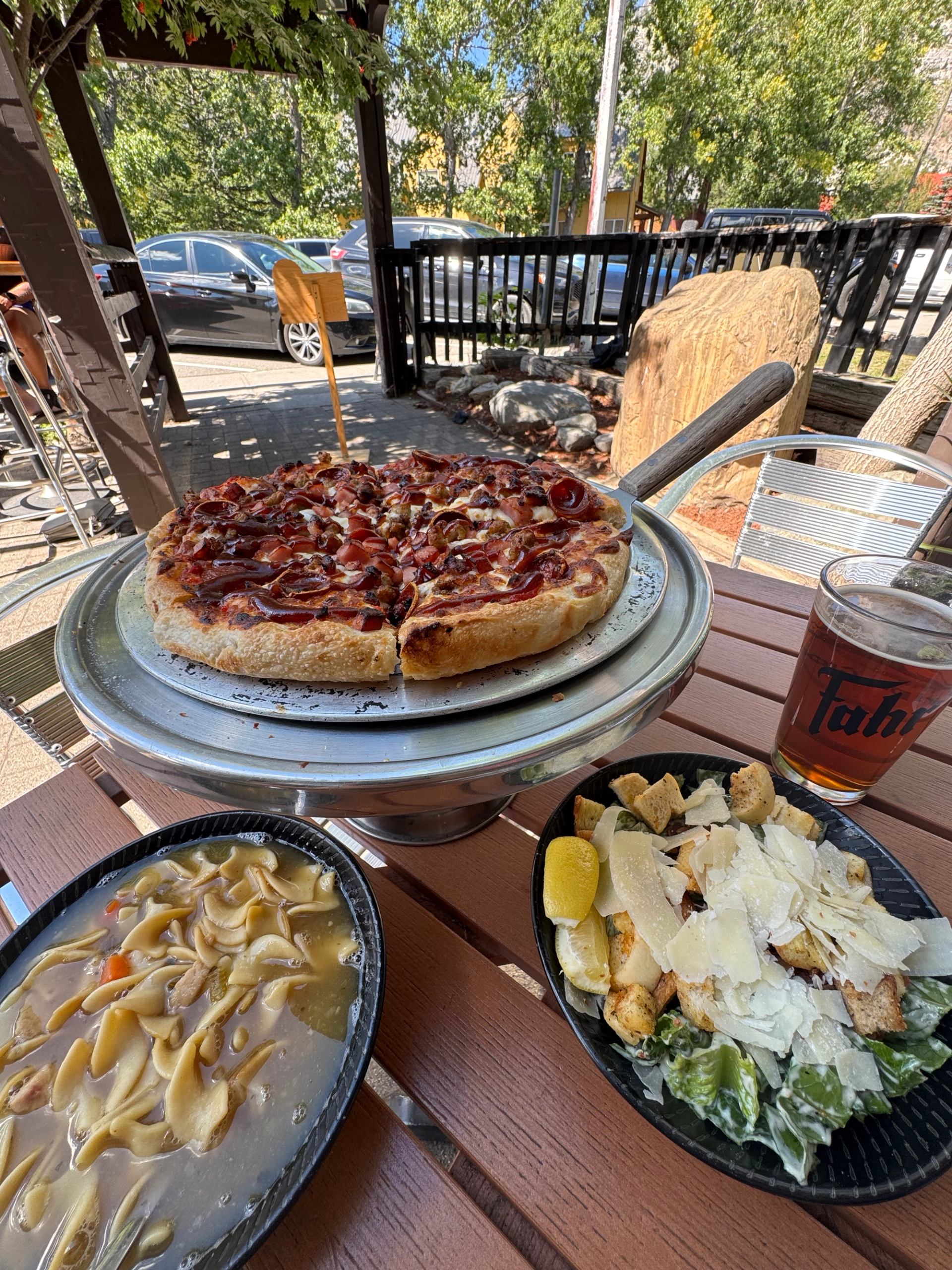 Deep-dish pizza, soup, Caesar salad, and iced tea on an outdoor dining table.