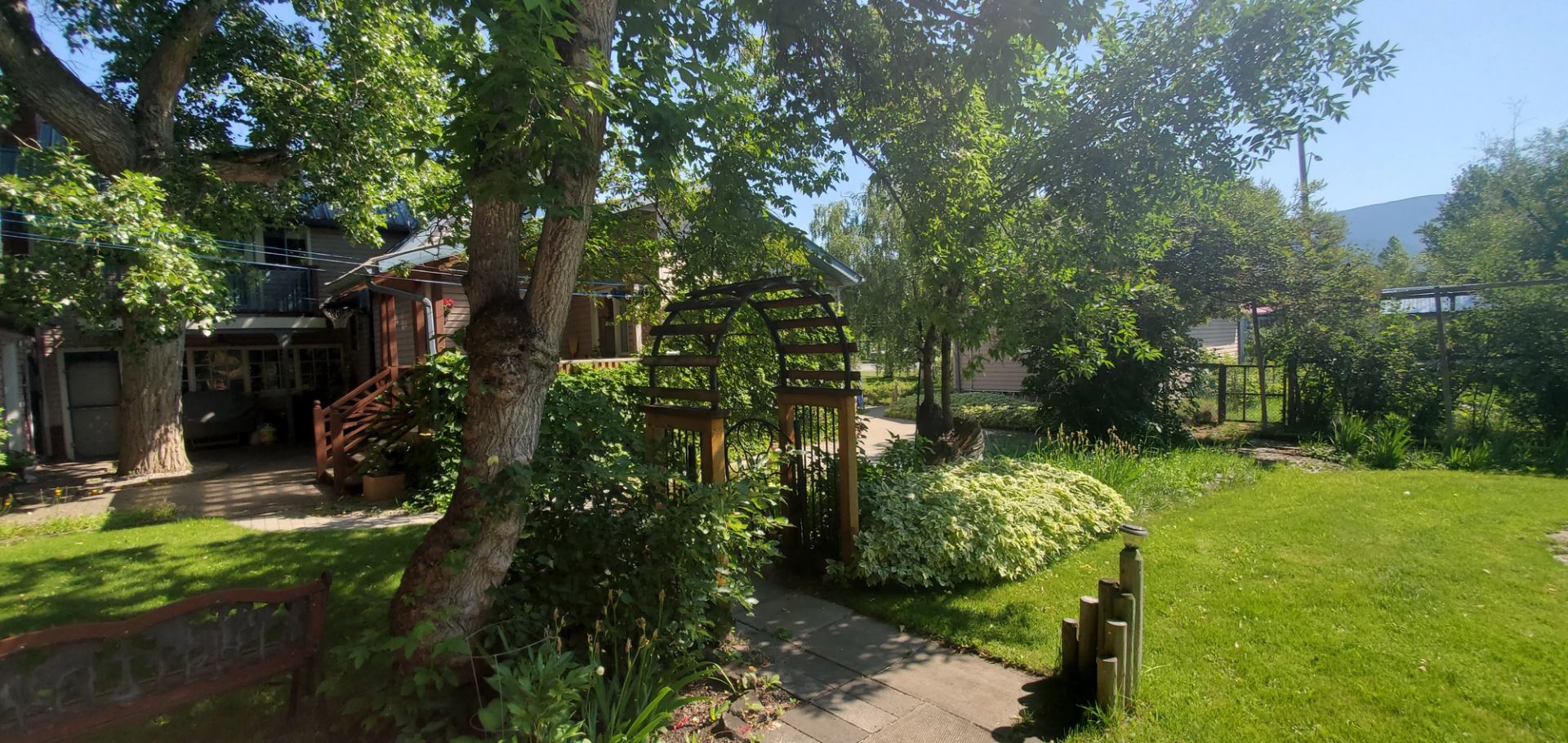 Garden path under wooden arbor beside lawn and trees, house in background.