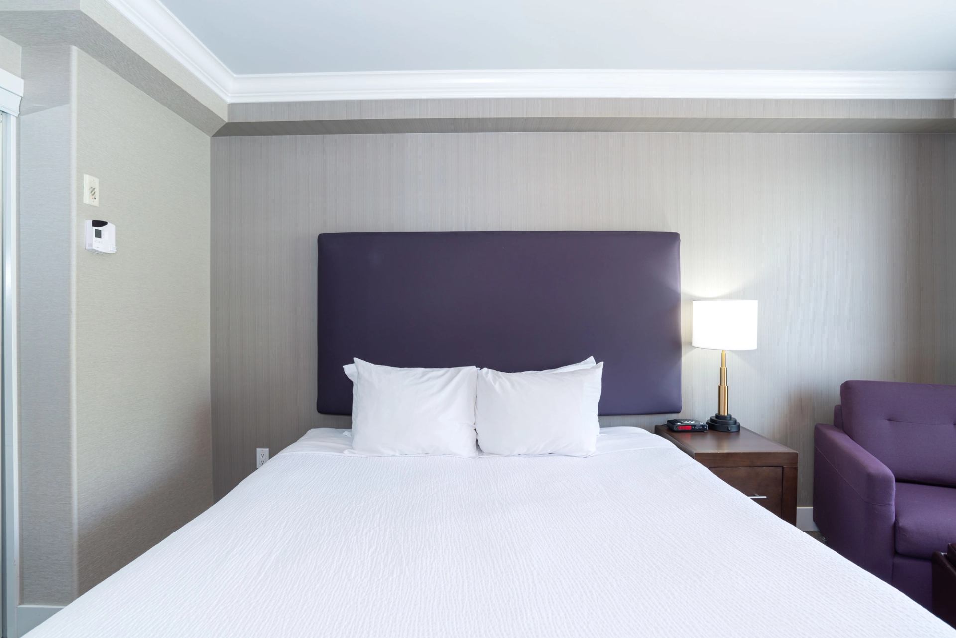 Hotel room bed with purple headboard, white bedding, nightstand lamp and purple chair.