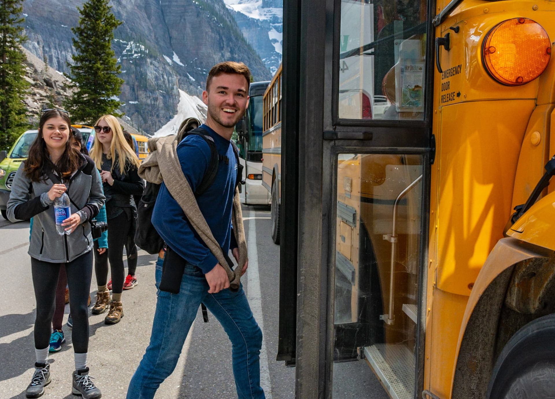 Smiling young man with a backpack stepping onto a yellow bus, with a group of people and snowy mountains in the background.