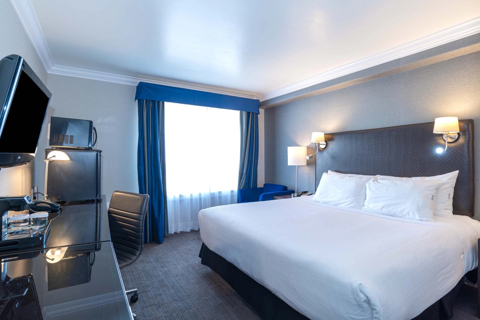 A hotel room with a king bed, work desk, television, and window with blue curtains.